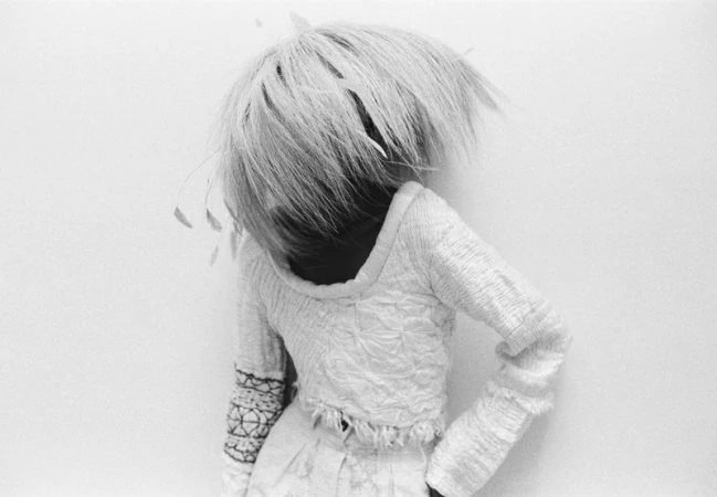 Person in textured white outfit with voluminous light hair against a plain white wall, black and white fashion photography.