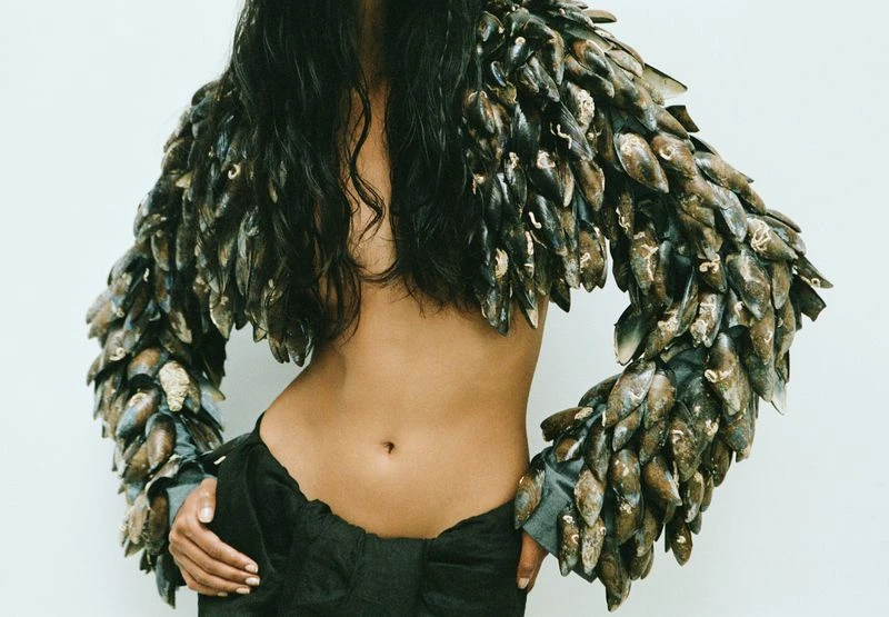 Adult female fashion model poses in a unique mussel-shell jacket and black skirt against a white background, showcasing avant-garde style.