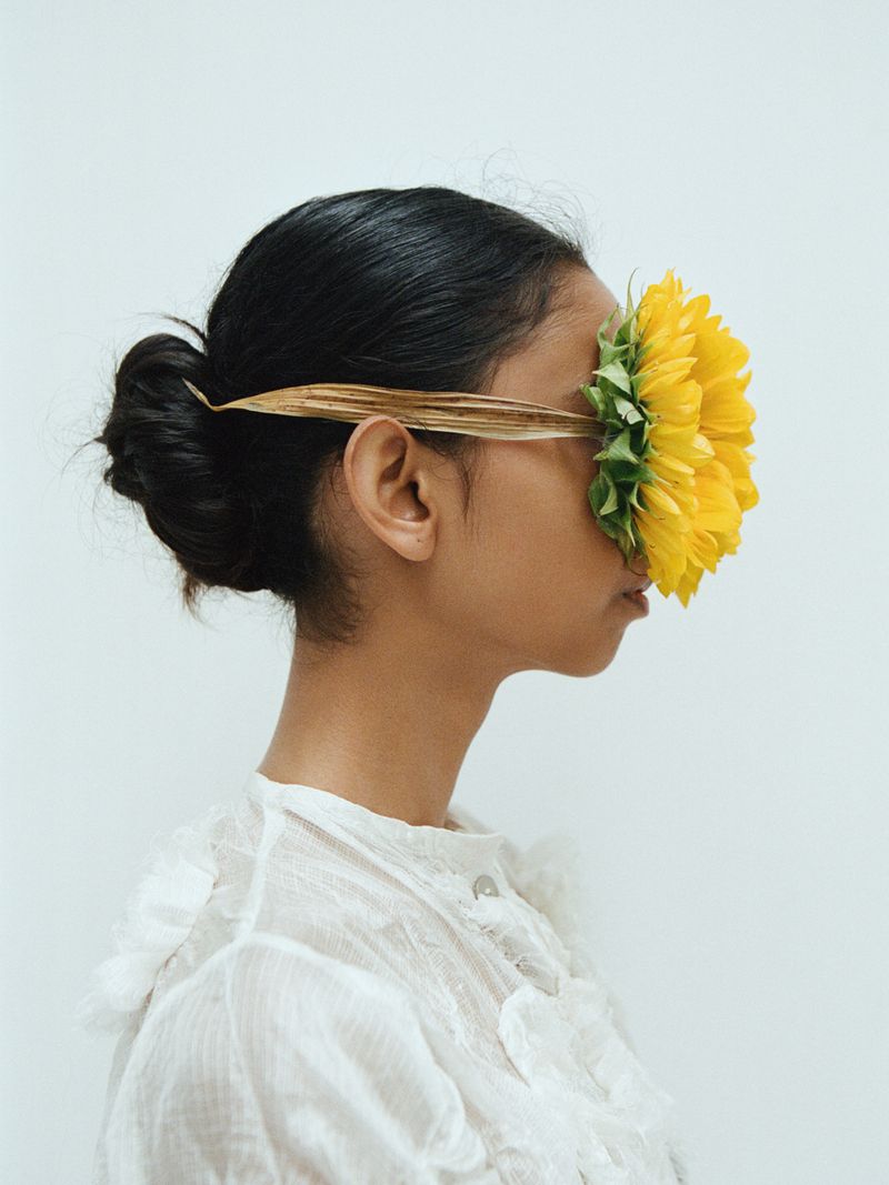 Profile of a person with dark hair and a white ruffled shirt, face partially obscured by a large bright yellow sunflower blossom.