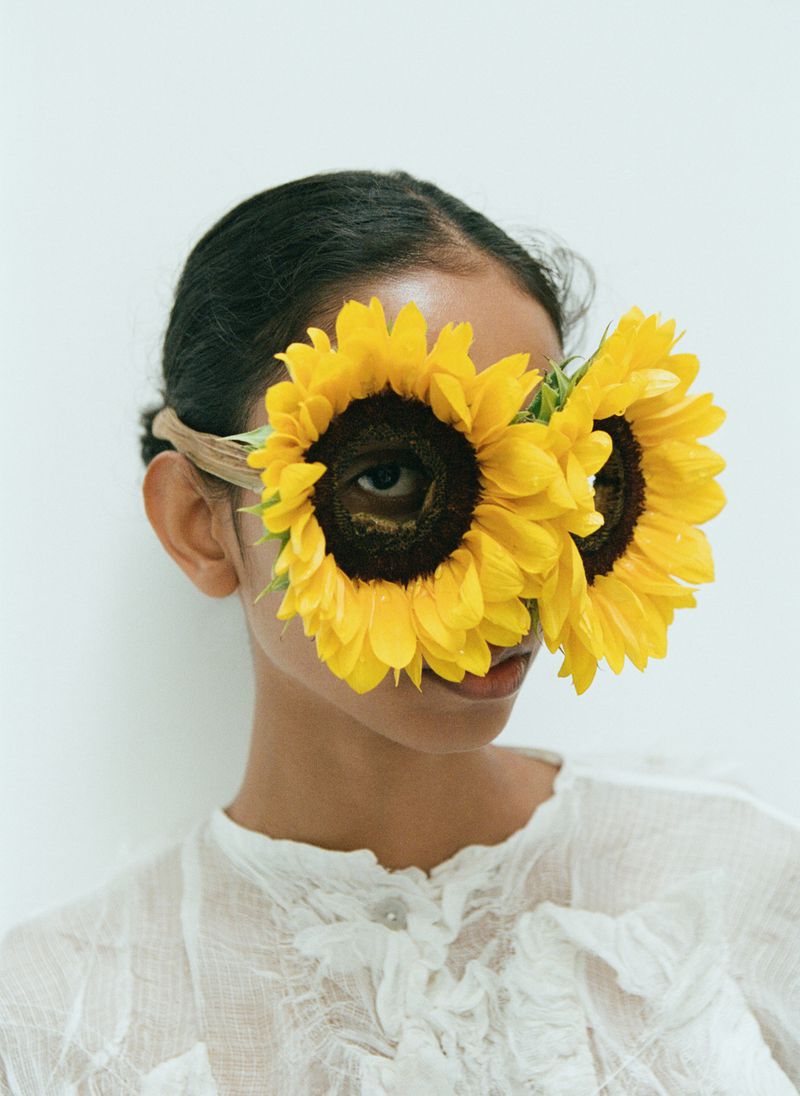 Woman with sunflowers covering her eyes in a studio portrait. The model is in a white top with a neutral background.