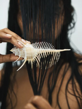 Conceptual beauty shot of a person holding a spiky conch shell in front of their face, obscuring it with dark, wet hair.