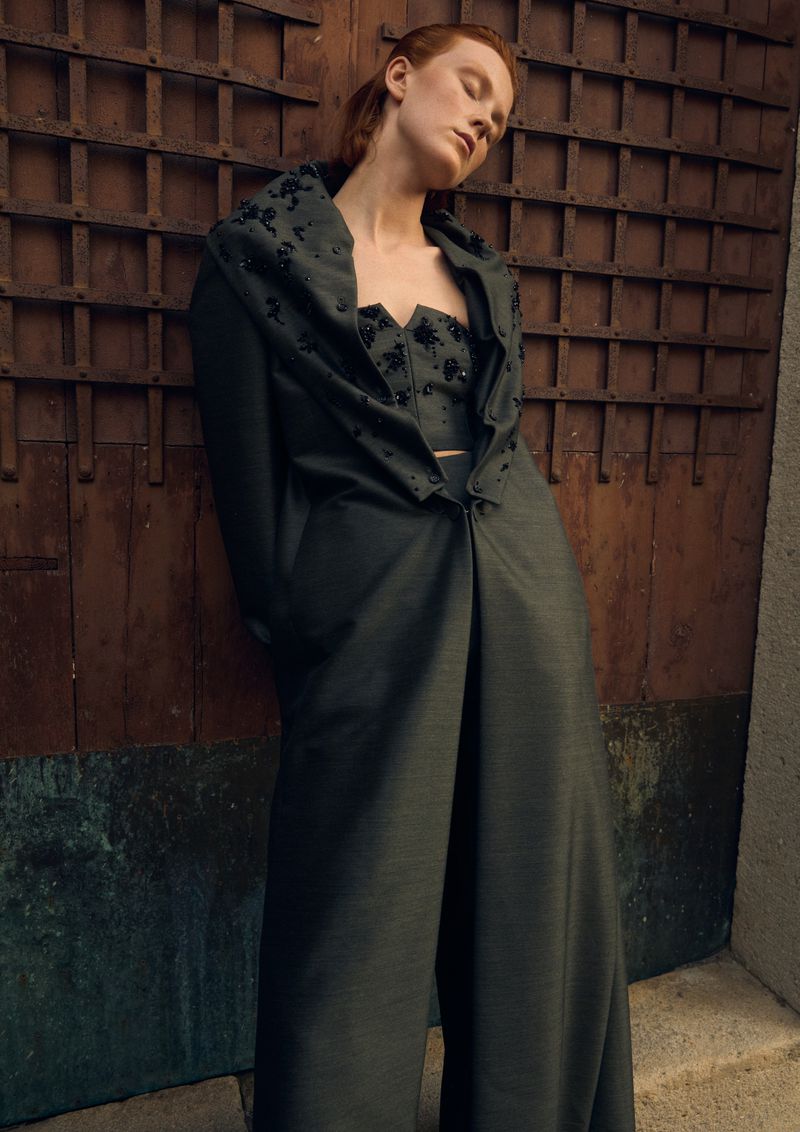 A serene, eye-level fashion shot of a redhead model in a dark green, embellished coat against a rustic wood door, bathed in soft light.