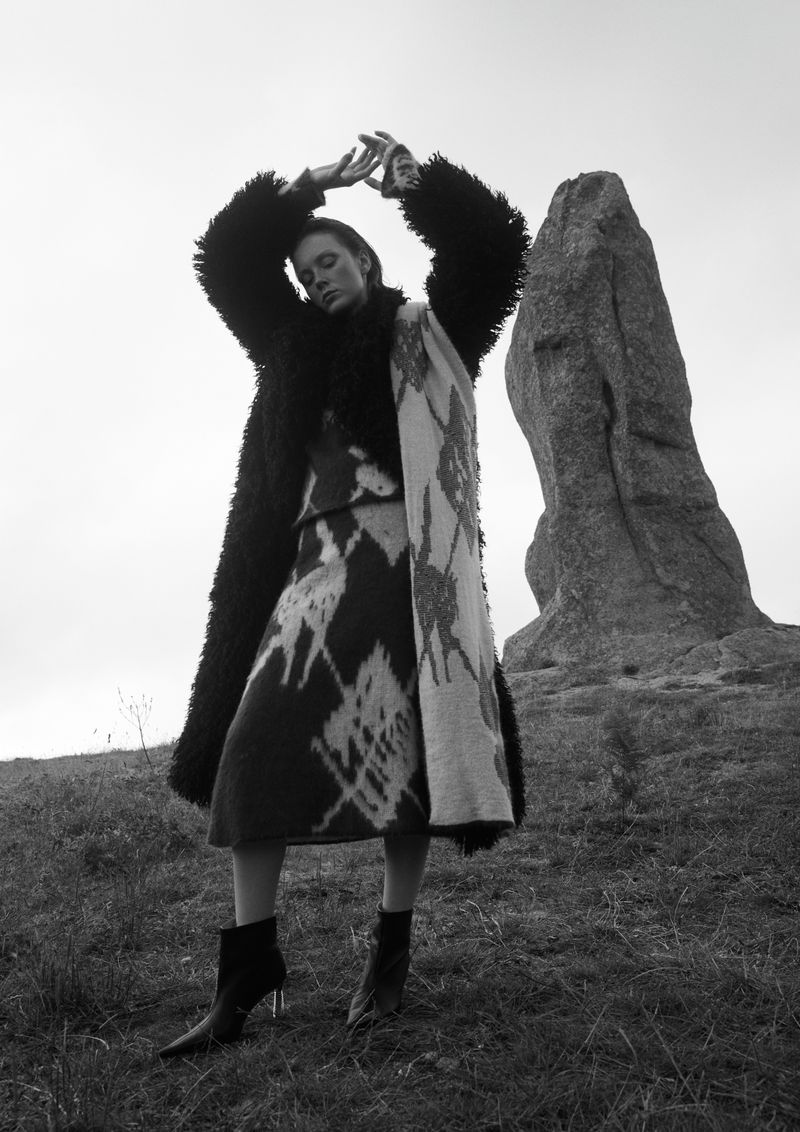 Woman in a fur coat and dress posing on grassy hill next to a rock formation under a bright sky in a black and white fashion shot.