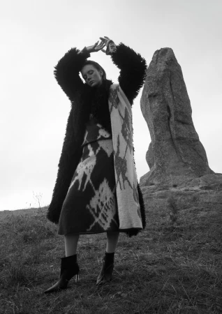 Woman in a fur coat and dress posing on grassy hill next to a rock formation under a bright sky in a black and white fashion shot.