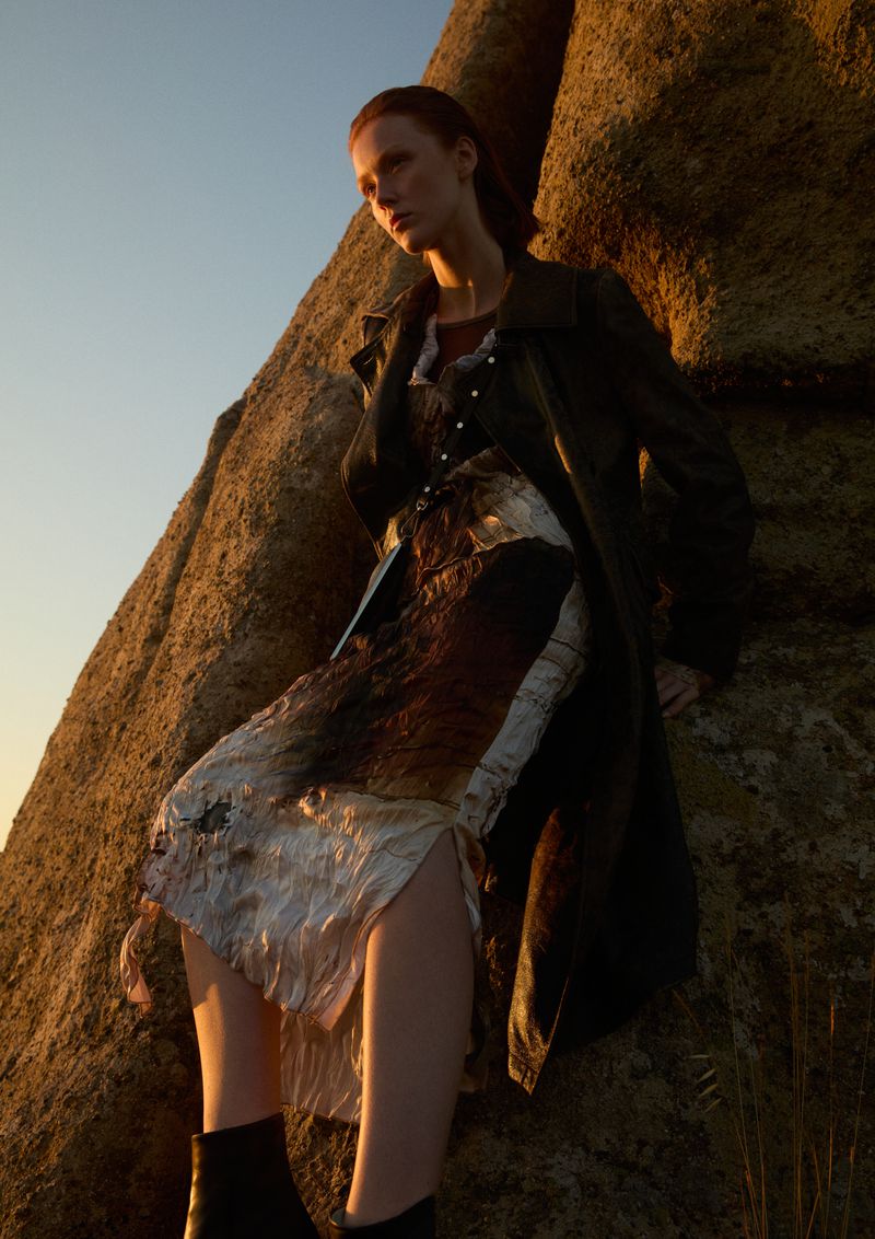 A red-haired woman in a patterned dress and black coat poses by a textured rock formation against a blue sky at golden hour.