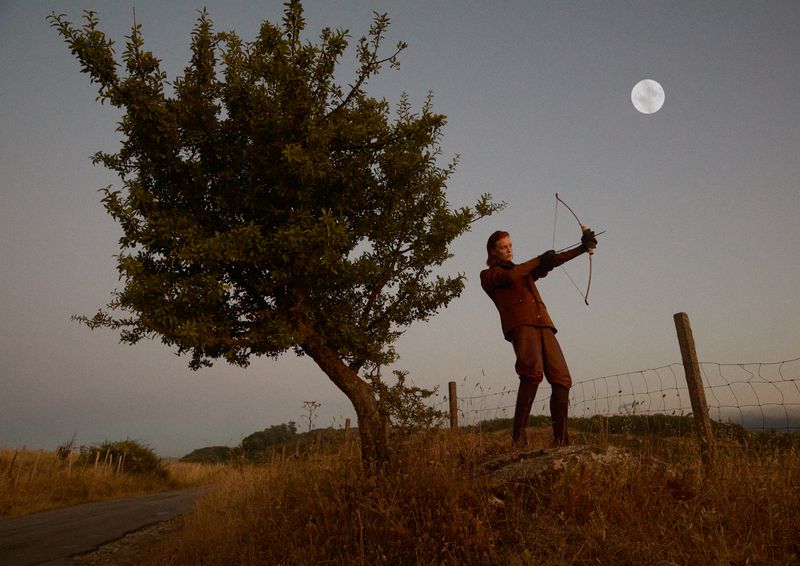 A person with a bow and arrow stands in a field by a tree, aiming towards the moon under a clear sky in a rural landscape.