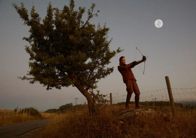 A person with a bow and arrow stands in a field by a tree, aiming towards the moon under a clear sky in a rural landscape.