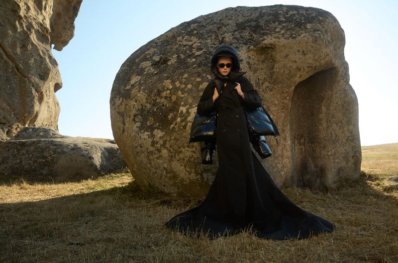 A female fashion model posing in a black dress and sunglasses, standing near a stone formation in a grassy field under a blue sky.