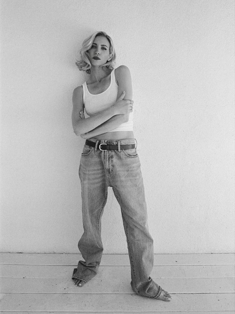 Black and white portrait of a woman in a white tank top and blue jeans standing barefoot indoors against a textured wall.