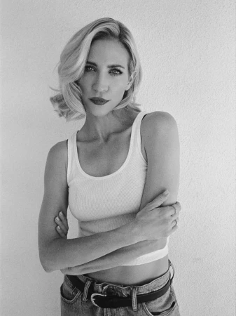 Portrait of an adult woman with blonde hair, wearing a white tank top and blue jeans, posing in front of a plain white wall. Black and white.