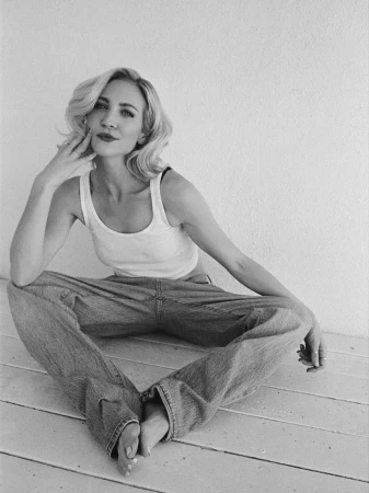 Black and white portrait of a blond woman with her hair in waves, sitting cross-legged against a white wall, wearing a tank top and jeans.