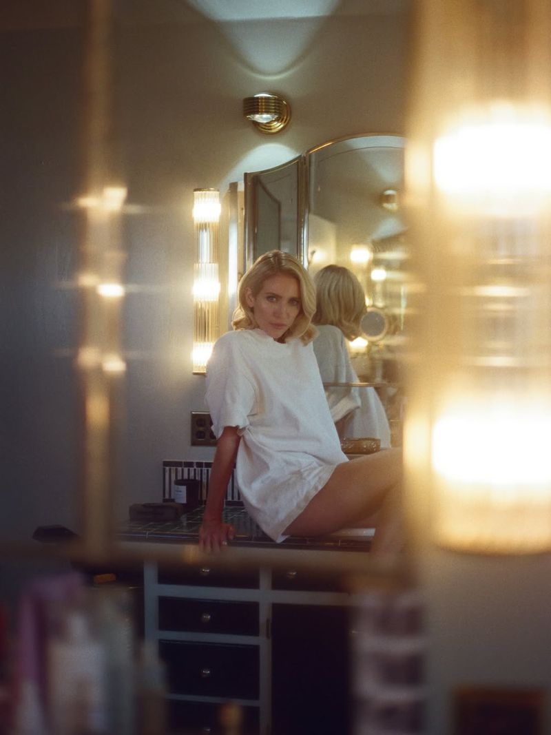 Blonde woman in a white shirt and shorts sitting on a vanity in a bathroom setting, with warm lighting and a reflective mirror.
