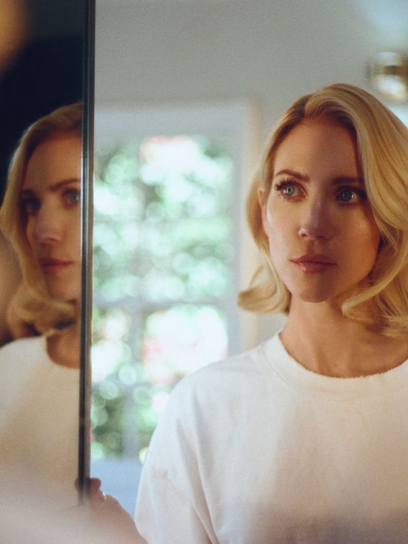 Blonde woman with shoulder-length waves gazes thoughtfully at her reflection in a mirror indoors with natural light.