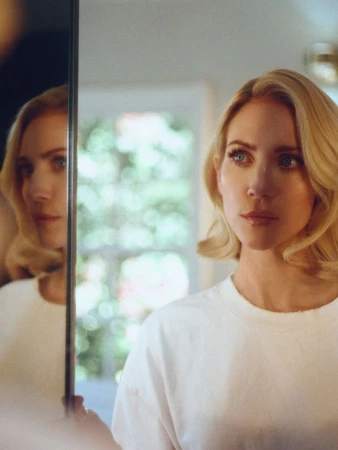 Blonde woman with shoulder-length waves gazes thoughtfully at her reflection in a mirror indoors with natural light.