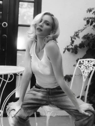 Brittany Snow seated on a white wrought-iron patio chair wearing a white tank top and jeans in black and white.
