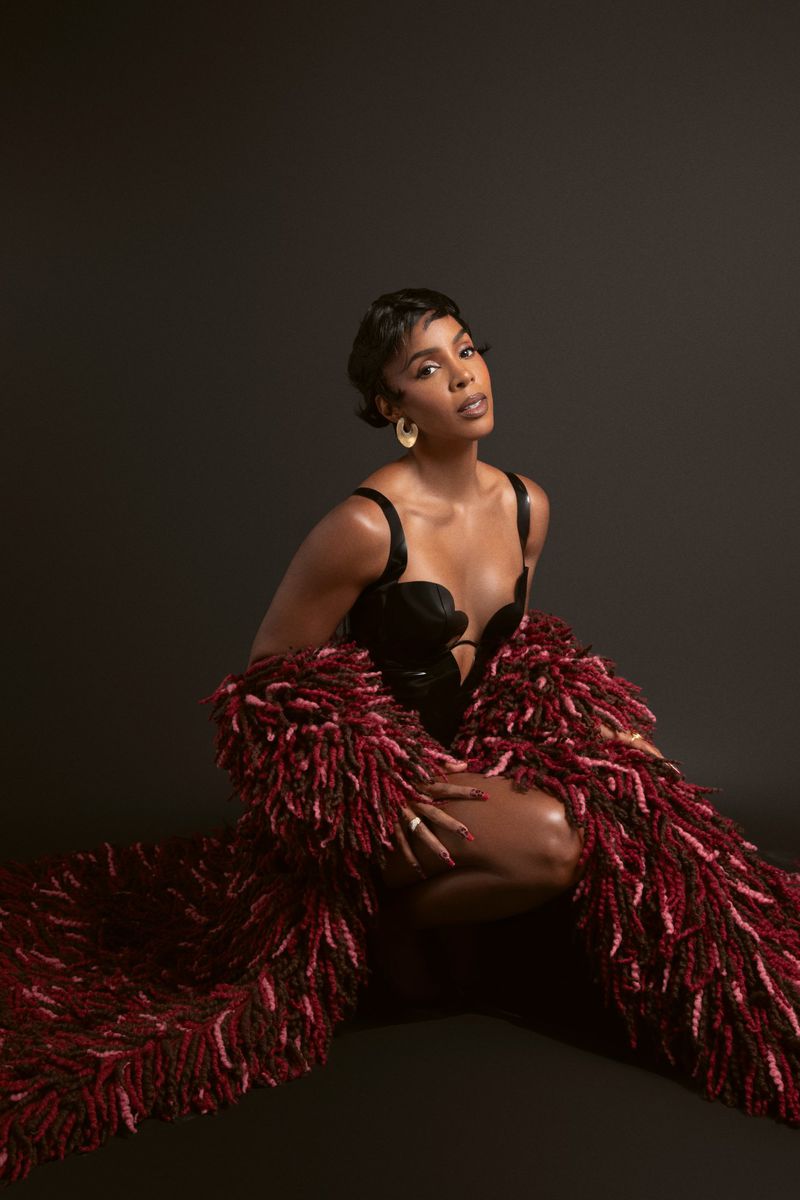 Portrait of a Black woman in a black corset top and a textured burgundy coat, kneeling in a photo studio against a dark background.