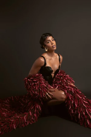 Kelly Rowland crouching in a black structured bodice and textured burgundy fringed coat against a dark studio background.