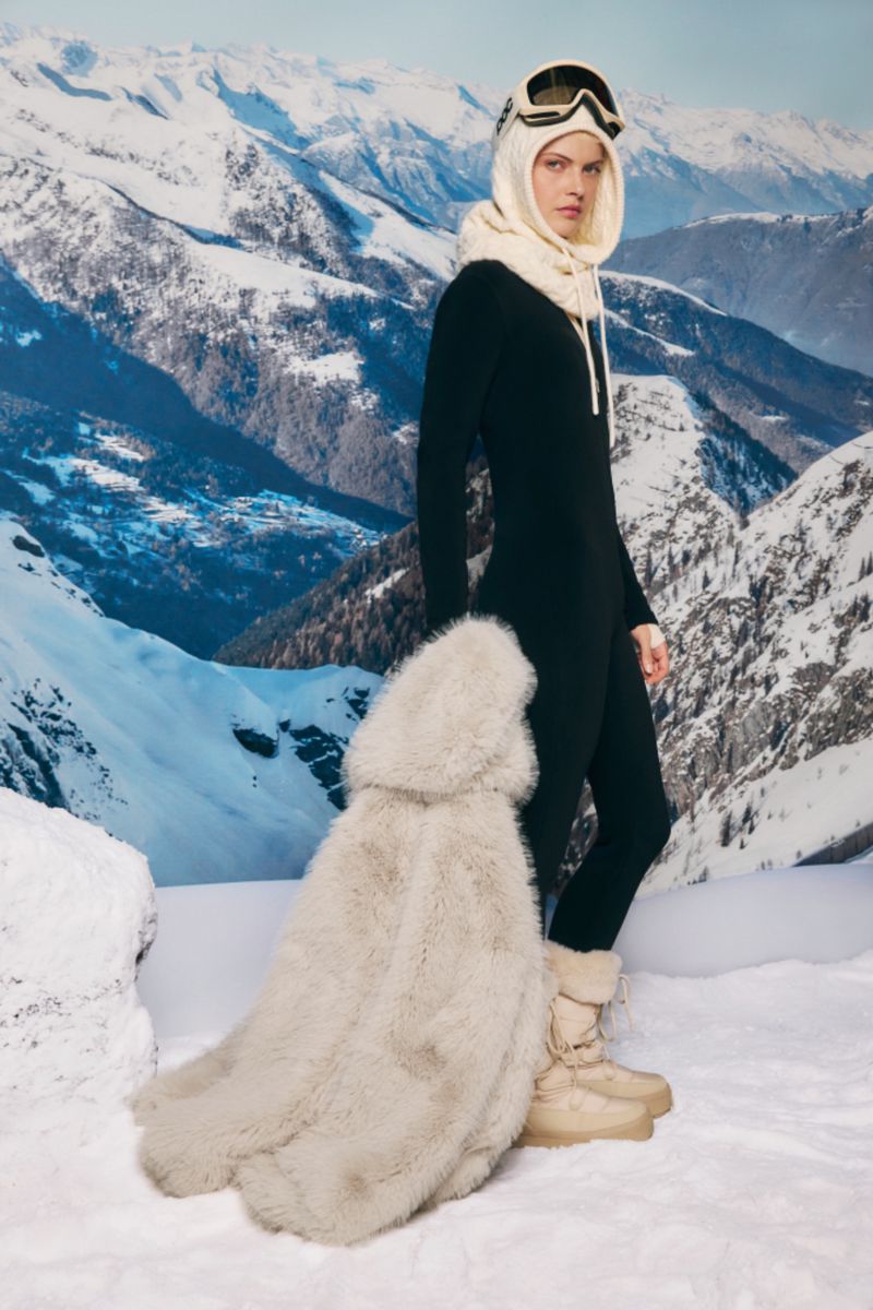 A woman in a black ski suit, winter boots, and goggles poses against a backdrop of snowy mountains, showcasing winter fashion.