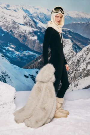A woman in a black ski suit, winter boots, and goggles poses against a backdrop of snowy mountains, showcasing winter fashion.