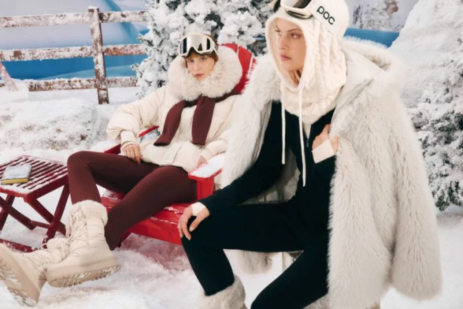 Two women wearing winter ski apparel sit on a red bench in a snow-covered outdoor setting. Ski goggles are visible. Winter fashion.