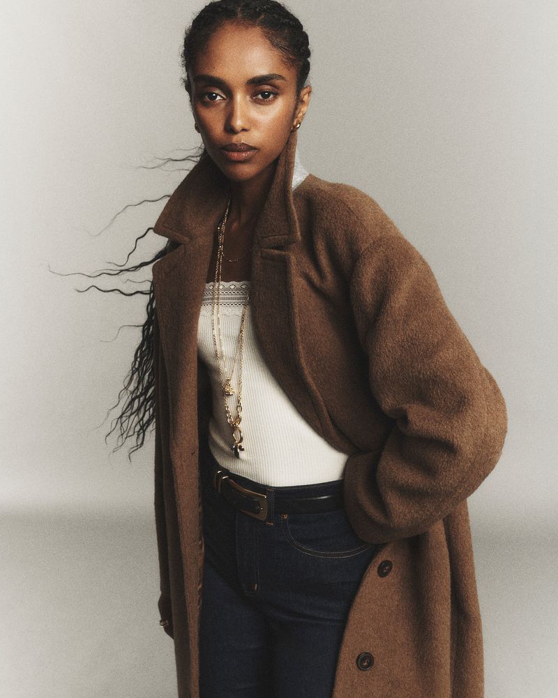 A woman with long hair wearing a brown coat over a white top and jeans, looking directly at the camera in a studio setting.