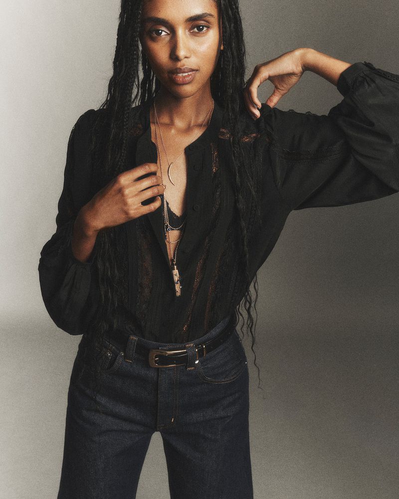 Elegant fashion portrait of a woman with long braids wearing a black blouse and blue jeans, posing in a neutral studio setting.