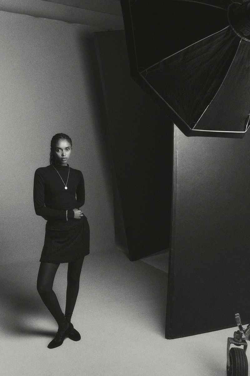 A woman stands confidently in a photo studio, wearing a black turtleneck dress, black stockings, and black shoes, looking straight at the camera.