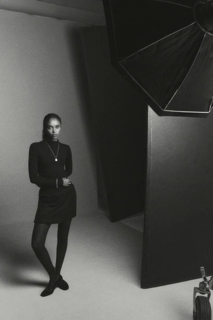 A woman stands confidently in a photo studio, wearing a black turtleneck dress, black stockings, and black shoes, looking straight at the camera.