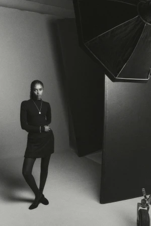 A woman stands confidently in a photo studio, wearing a black turtleneck dress, black stockings, and black shoes, looking straight at the camera.