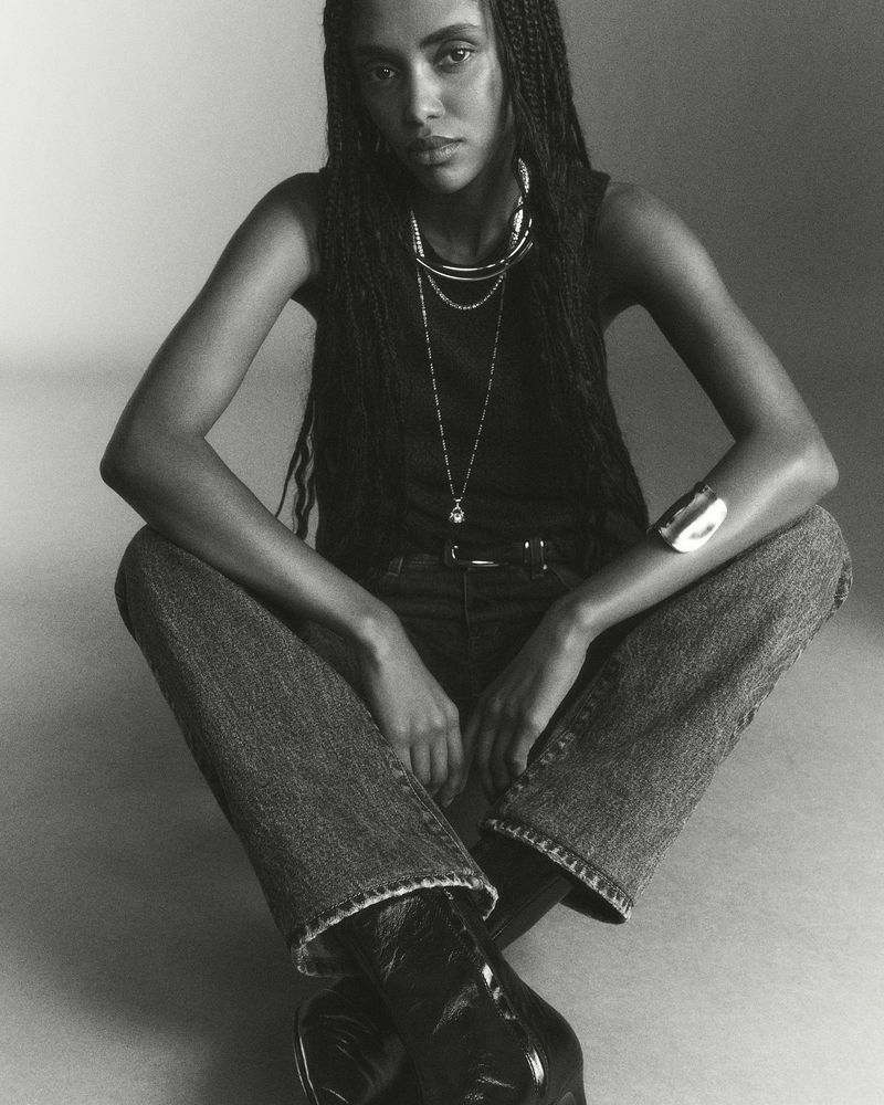 Black and white portrait of a woman in a tank top, jeans, and boots, seated with layered necklaces and a bracelet, indoor fashion shot.