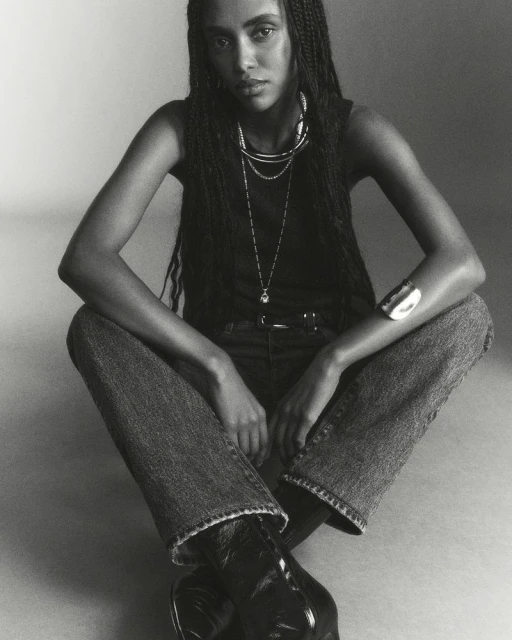 Black and white portrait of a woman in a tank top, jeans, and boots, seated with layered necklaces and a bracelet, indoor fashion shot.