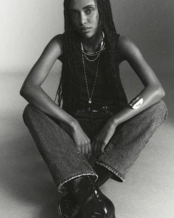 Black and white portrait of a woman in a tank top, jeans, and boots, seated with layered necklaces and a bracelet, indoor fashion shot.