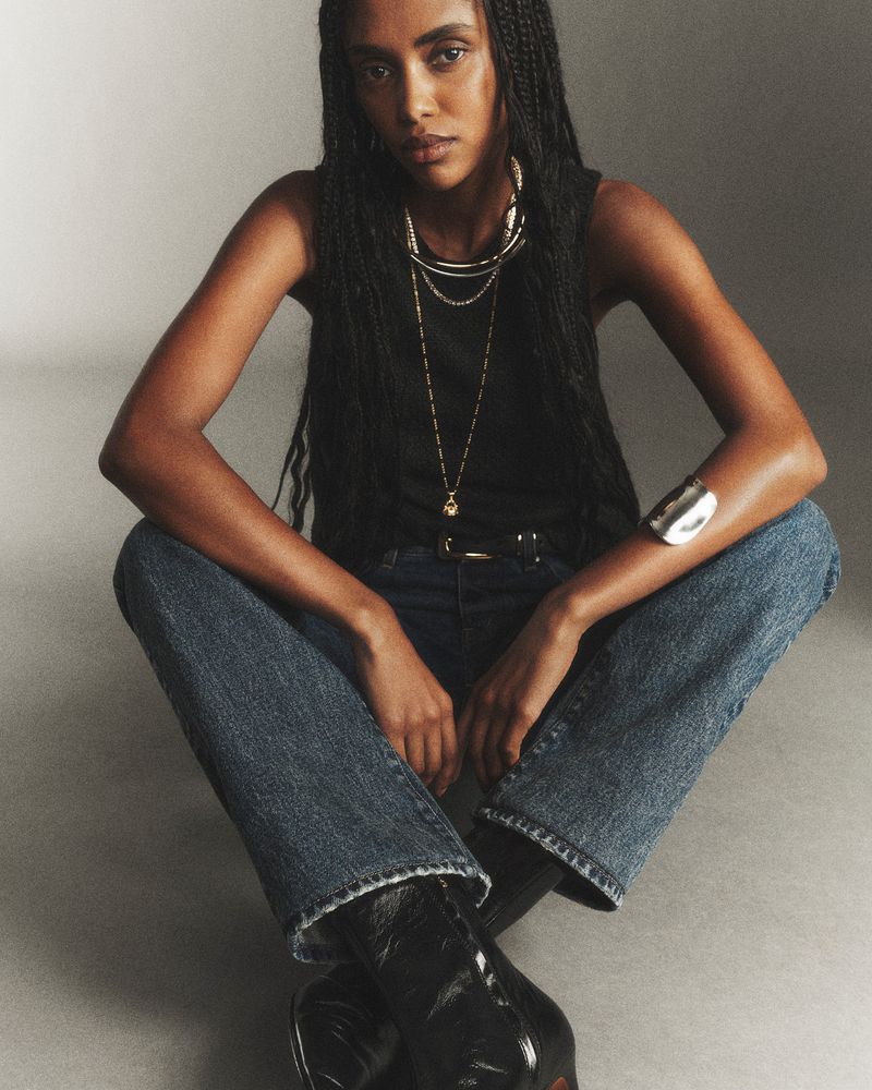 A dark-skinned woman poses in a black top, jeans, and boots against a neutral backdrop, showcasing jewelry and a bracelet.