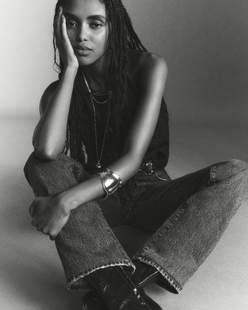 Monochrome portrait of a young woman with braided hair, wearing casual jeans and a dark tank top, seated against a plain background.