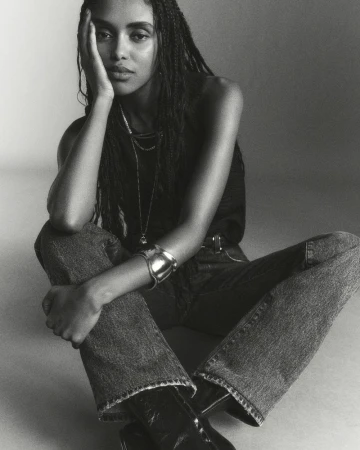 Monochrome portrait of a young woman with braided hair, wearing casual jeans and a dark tank top, seated against a plain background.