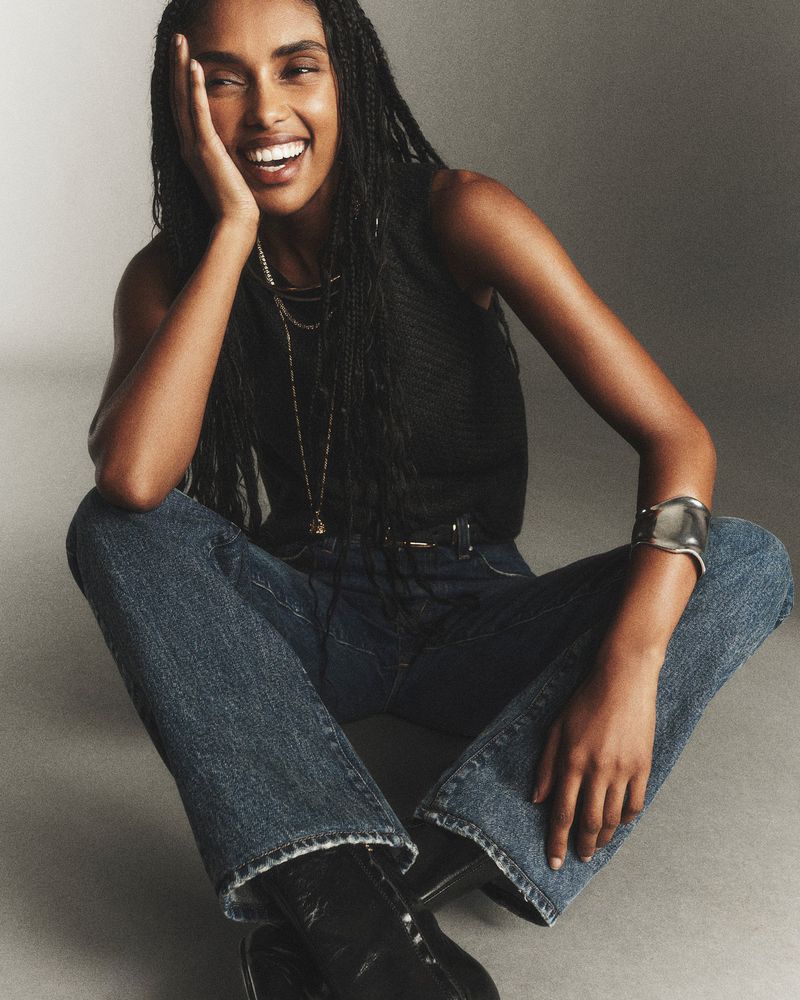 Smiling dark-skinned woman with braided hair, wearing jeans and a black top, poses in a casual setting with natural light.