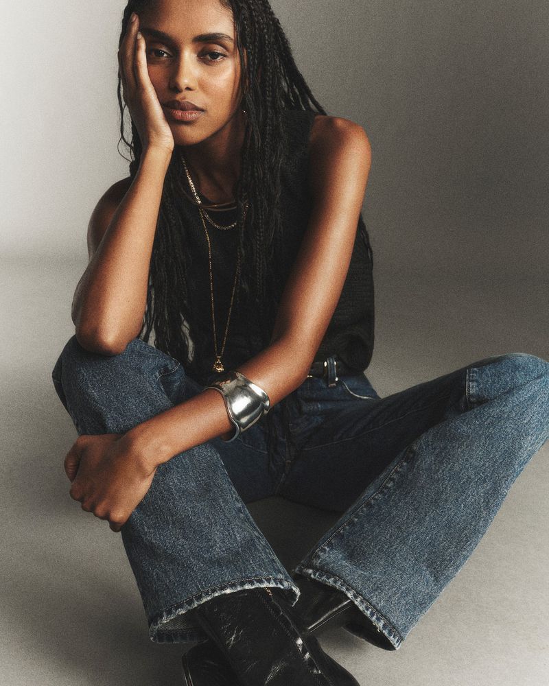 An adult female sits on the floor with her hand near her face, wearing a black top, blue jeans, jewelry and black boots in a studio.