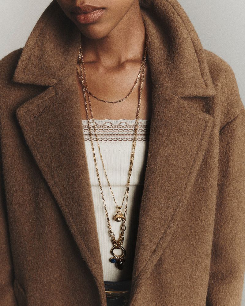 Close-up of a black woman wearing a brown wool coat and layers of gold necklaces, showcasing fashion and style in a studio setting.