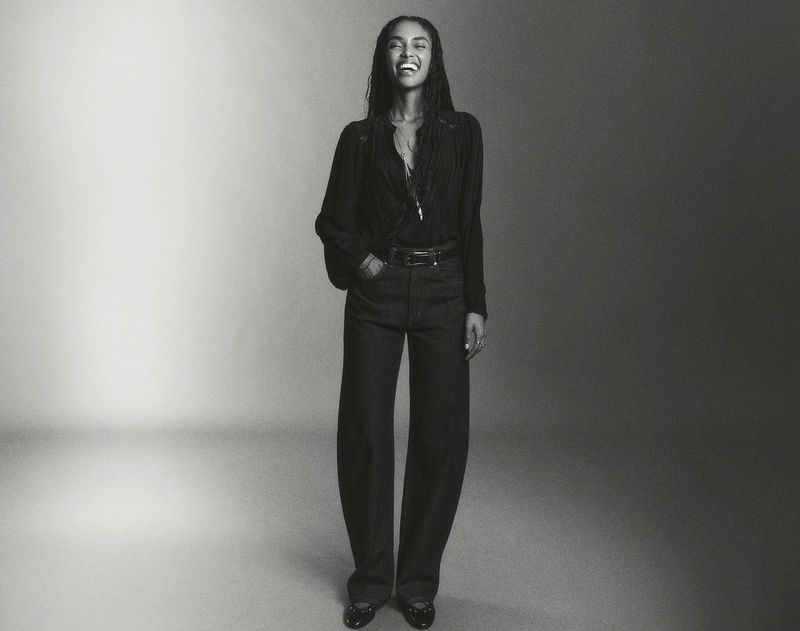 Full-length portrait of a smiling adult woman with dark hair, wearing a long-sleeved top and wide-leg jeans, posed against a gradient grey background.