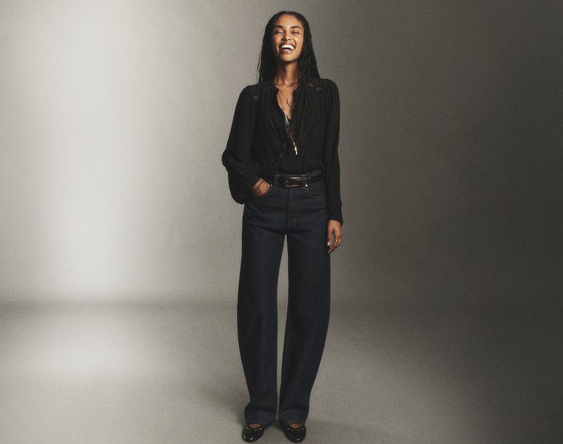 A full-length shot of a woman in a black blouse and jeans, standing with a joyful expression against a gradient gray background.
