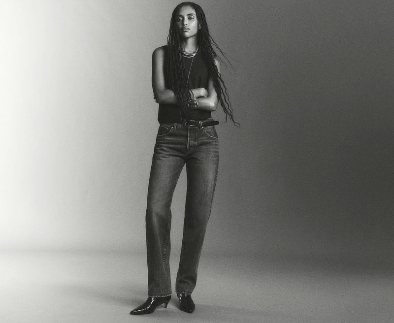 A stylish adult female is posing indoors wearing a black top, denim jeans, a black belt, and pointed-toe heels in a studio setting.