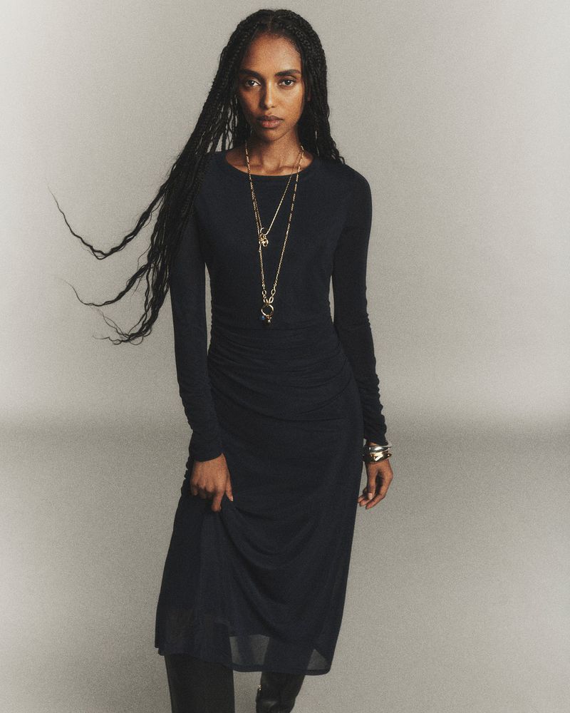 A woman with long dark hair is standing in a studio setting, wearing a long-sleeved black dress and jewelry, posing for a fashion shot.