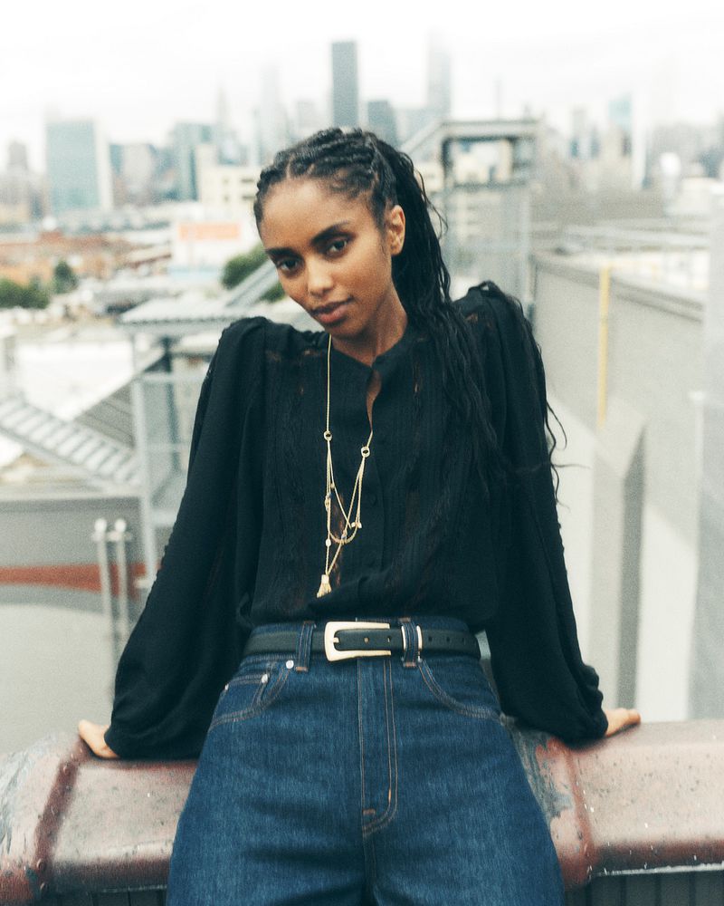 Adult female posing on rooftop with skyline background wearing jeans, black blouse, and gold necklace in outdoor setting.