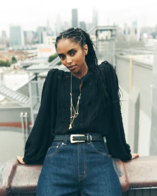 Adult female posing on rooftop with skyline background wearing jeans, black blouse, and gold necklace in outdoor setting.