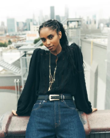 Adult female posing on rooftop with skyline background wearing jeans, black blouse, and gold necklace in outdoor setting.
