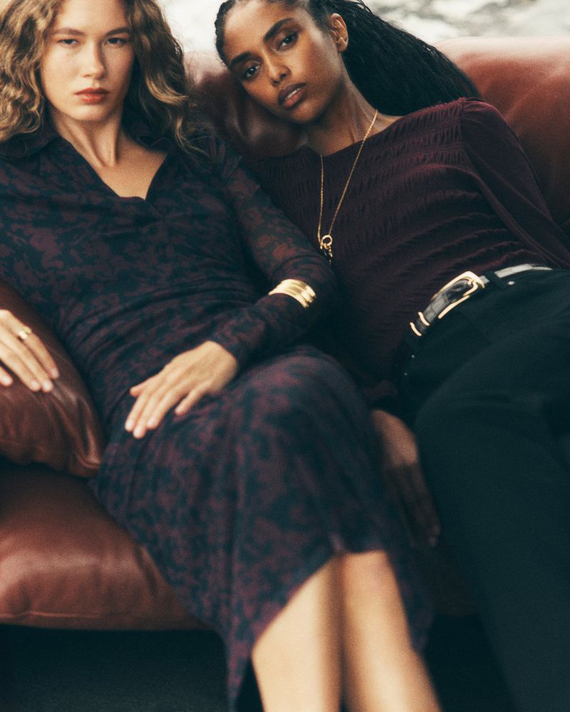 Two women pose on a brown leather sofa. One woman wears a printed long-sleeve dress, while the other wears a dark top and black pants.
