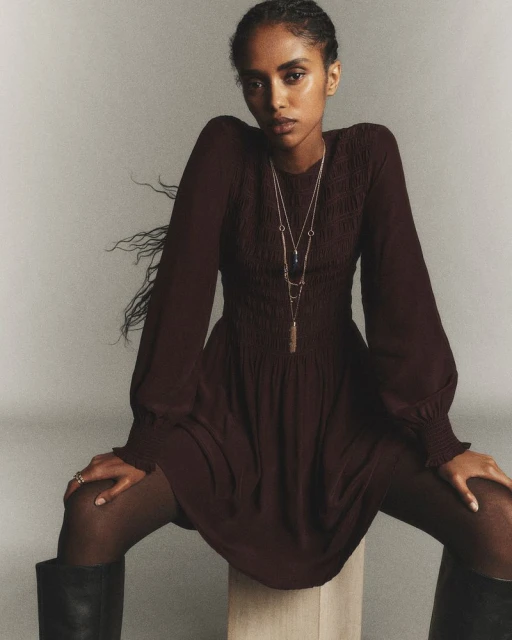 Dark-haired woman poses in a long-sleeve dark-burgundy dress, sheer tights, and high boots in a studio with a neutral backdrop.