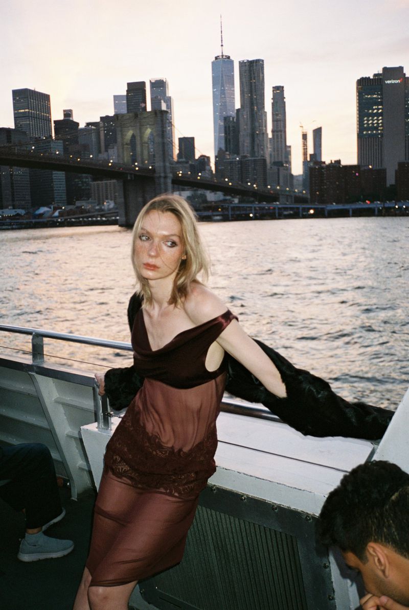 A young woman poses on a boat with a sheer brown dress and black jacket, against the backdrop of the Manhattan skyline and Brooklyn Bridge.