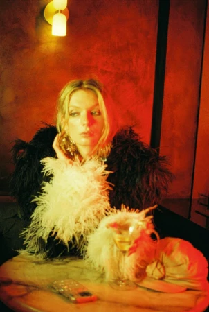 Model Willa Devereux wearing a black and white feather coat seated at a marble table under warm red lighting.