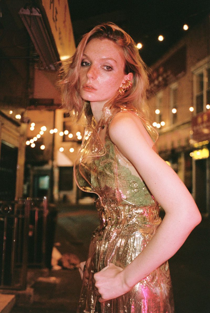 Portrait of an adult woman in a transparent and gold dress posing on a city street at night, illuminated by string lights.